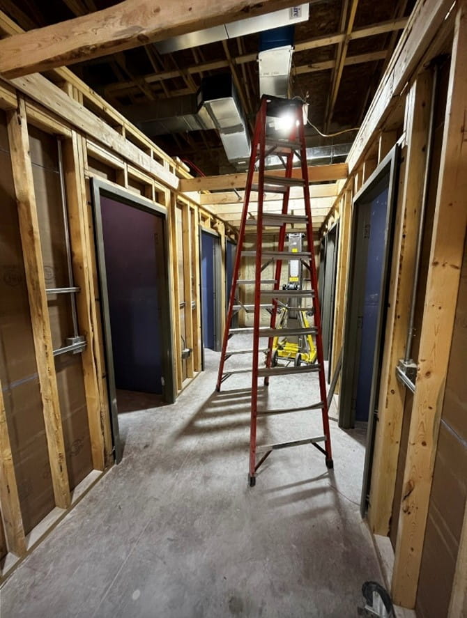 A room hallway with exposed walls and ceiling, a concrete floor, and doorways on either side. There is a ladder in the middle of the hallway under a light fixture. 