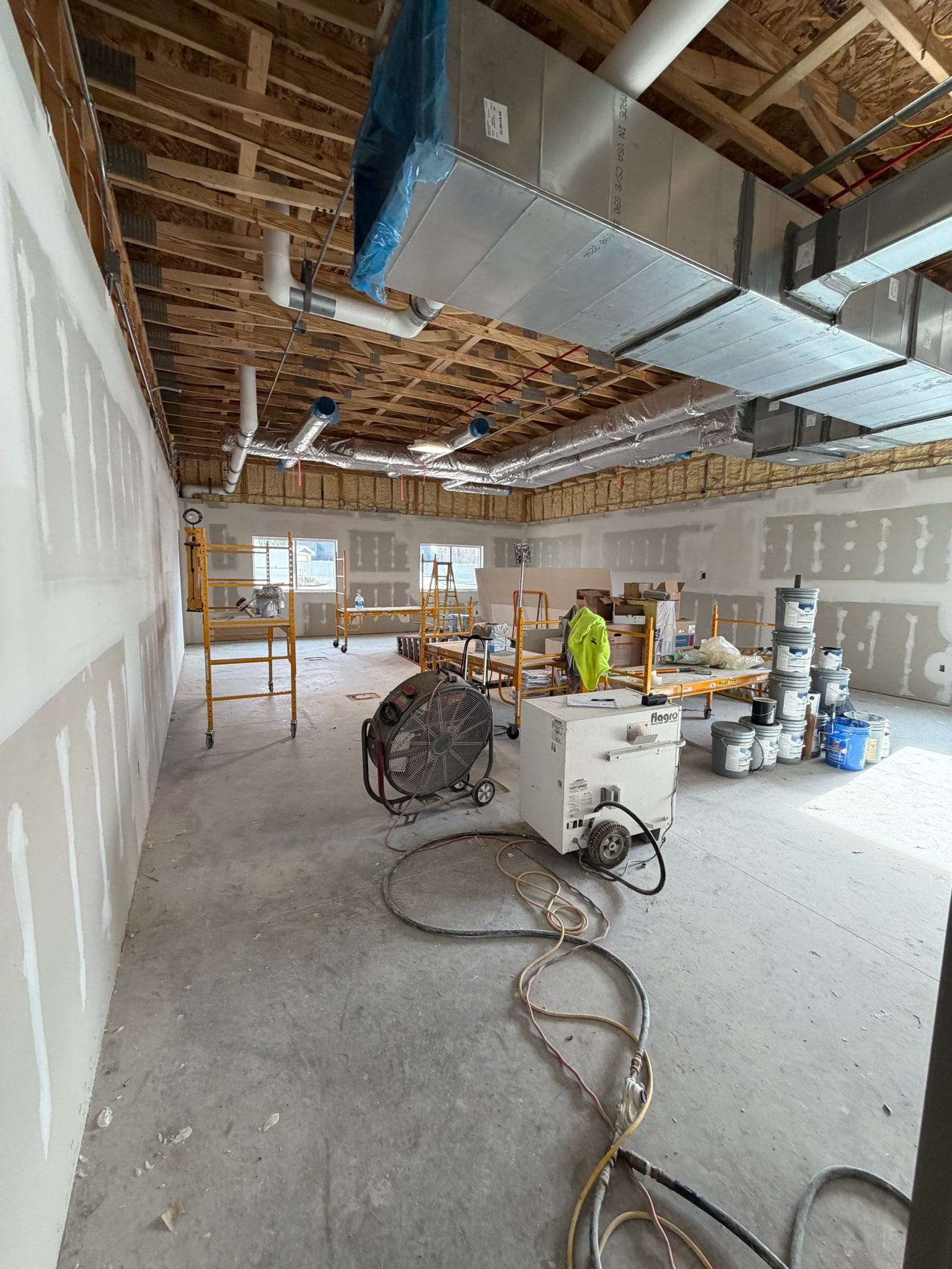 The south classroom with new drywall, unfinished floor, and exposed ceiling, There is construction equipment in the middle of the room.