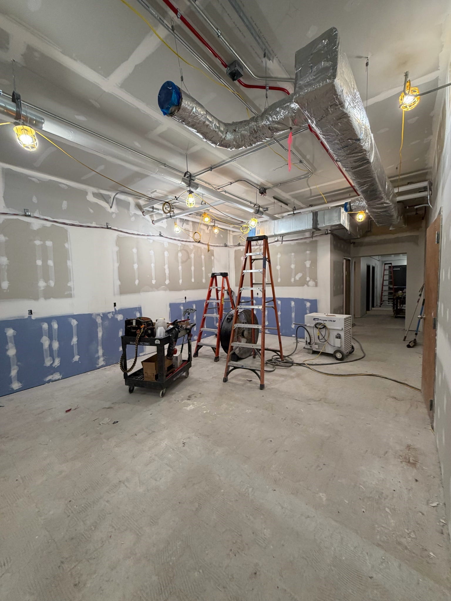 The student break room with new drywall and unfinished floor. There are ladders and construction equipment in the middle of the room.