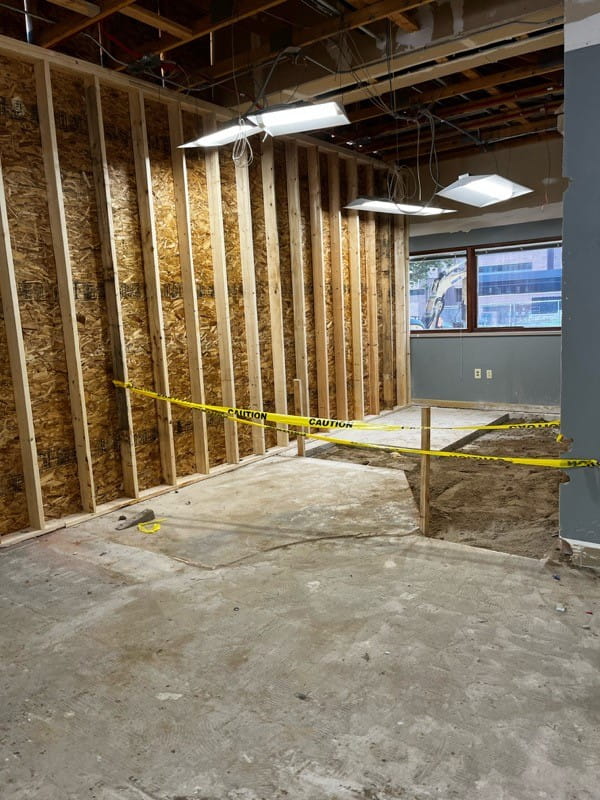 an unfinished room with exposed studs, exposed ceiling and cement floor and caution tape around an area cut out of the floor where dirt is exposed