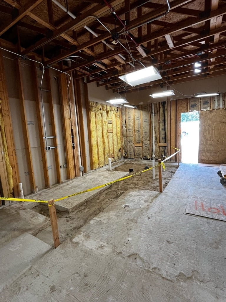 an unfinished room with exposed studs, exposed ceiling and cement floor and caution tape around an area cut out of the floor where dirt is exposed