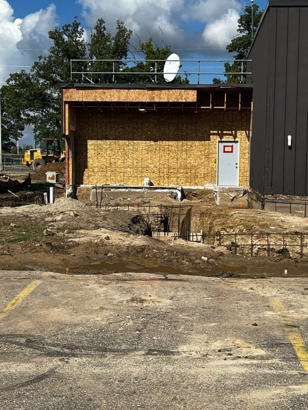 A dug up area with exposed plumbing and rebar in front of a building with a grey door surrounded by a plywood wall.