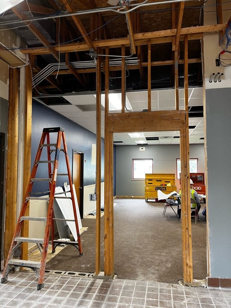 A doorway frame with the wall removed and a ladder standing where a wall used to be. Inside the room are tool chests and construction equipment.