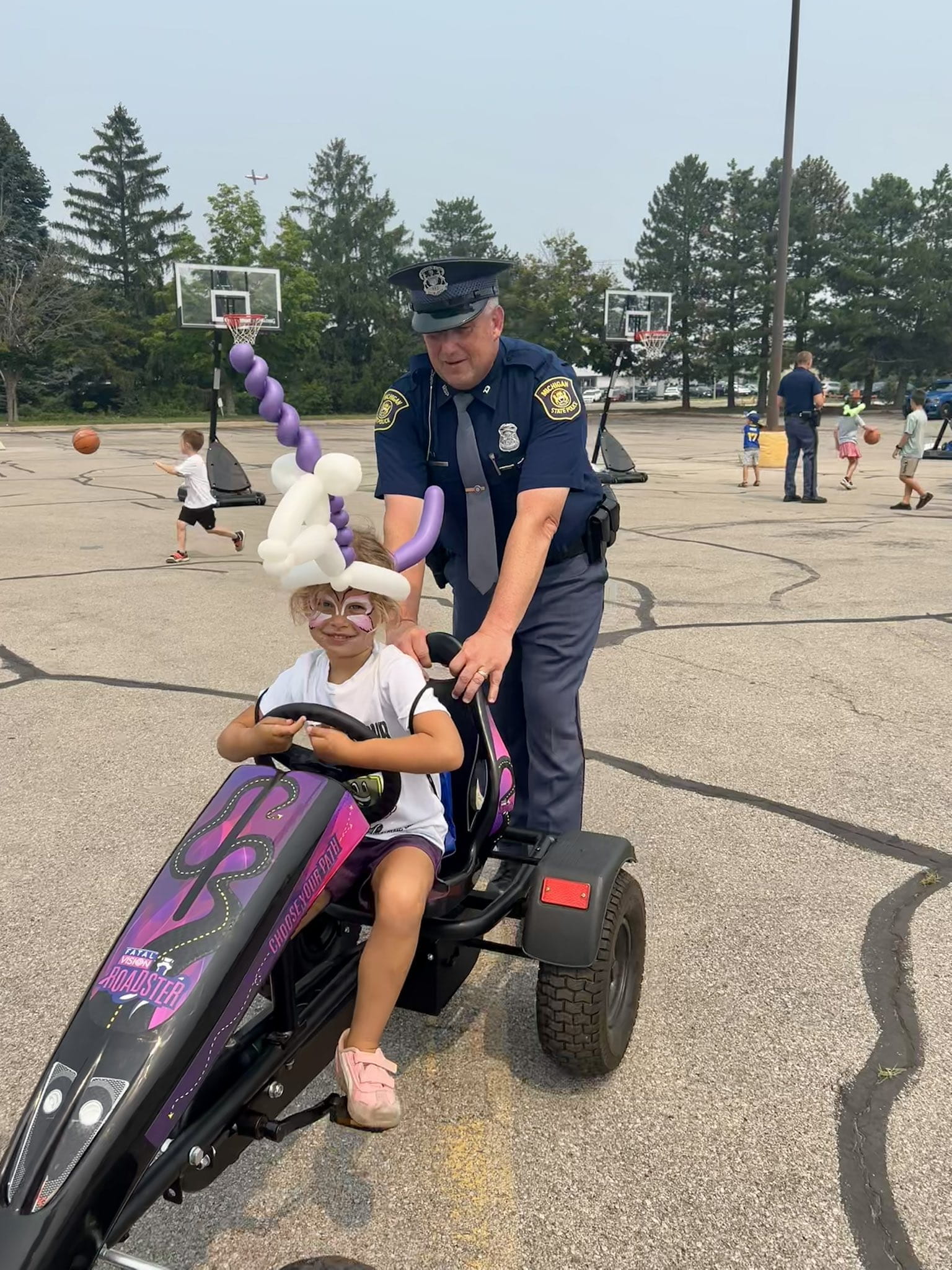 MSP trooper pushing a child on a tricycle with basketball court in background. 