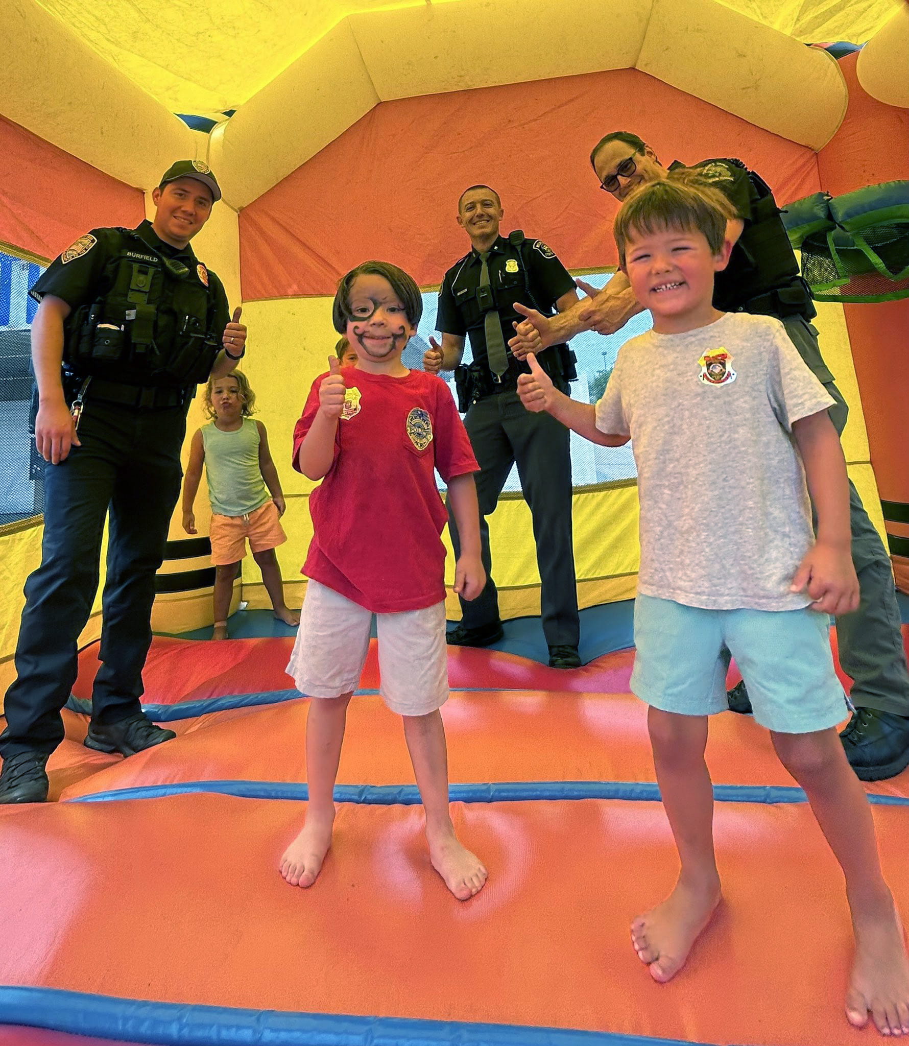 Police officers and children in a bounce house giving thumbs up hand signs. 