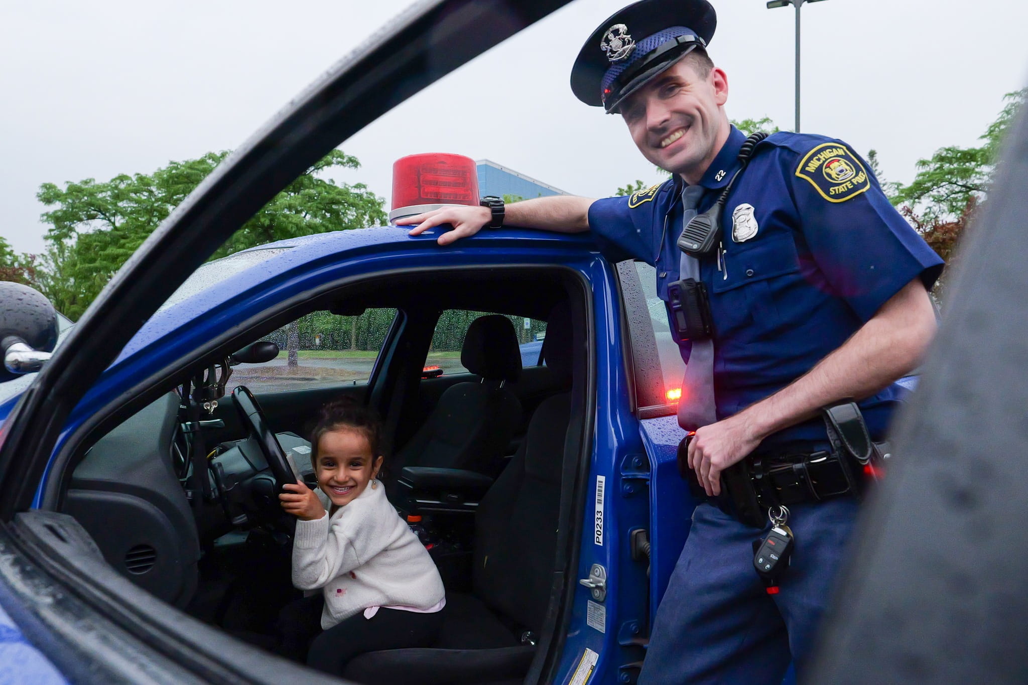 Young child sitting in MSP patrol car with a trooper standing near the open door. 
