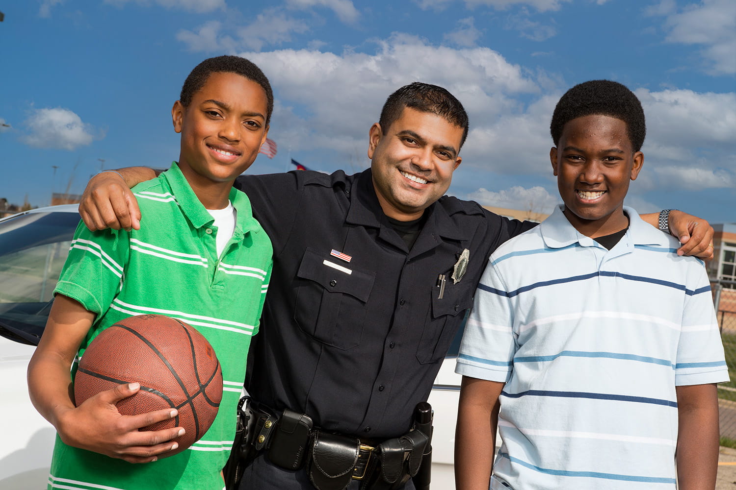 Latino school resource officer with 2 African-American teen boys holding a basketball outside of school. 