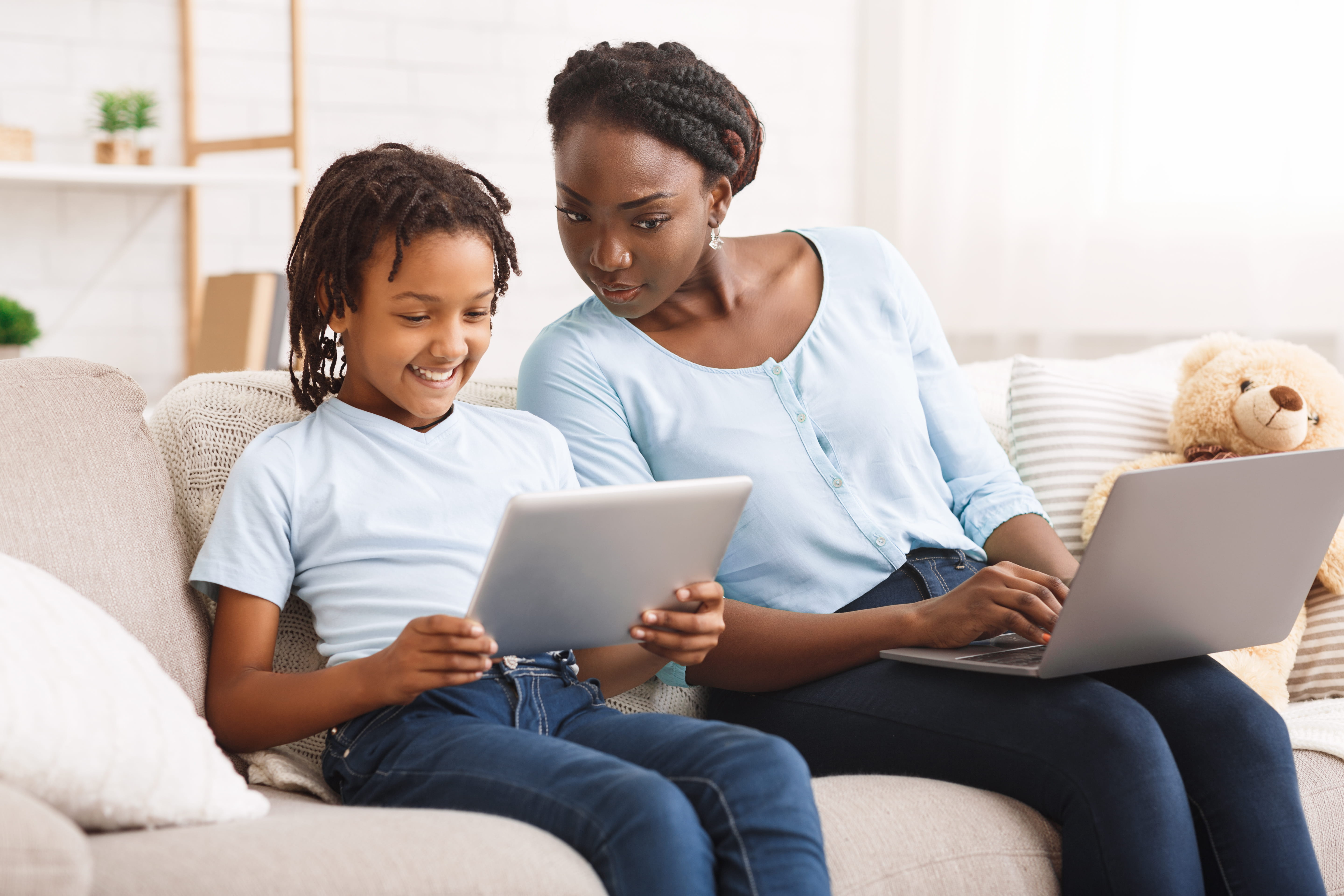 Young girl on tablet with mother sitting next to her on laptop, observing girl's online activity. 