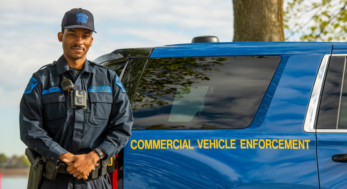 Male motor carrier officer standing in front of a Michigan State Police Commercial Vehicle Enforcement Division patrol car. 