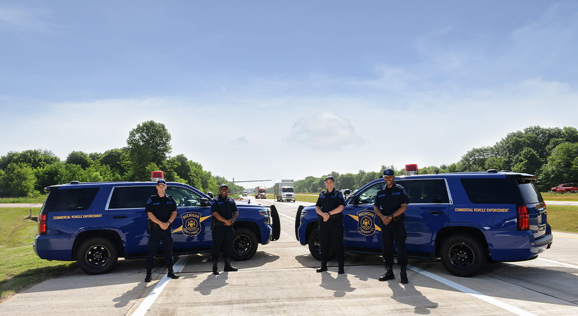 Four motor carrier officers stand in front of patrol vehicles on the road. 