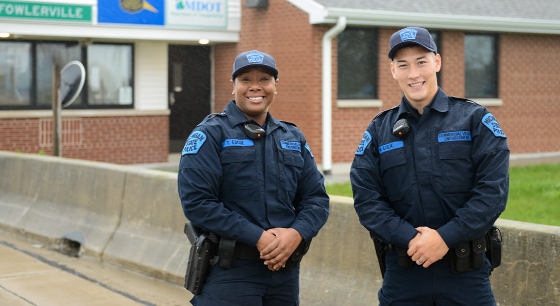 Two motor carrier officers stand in front of a weigh station. 