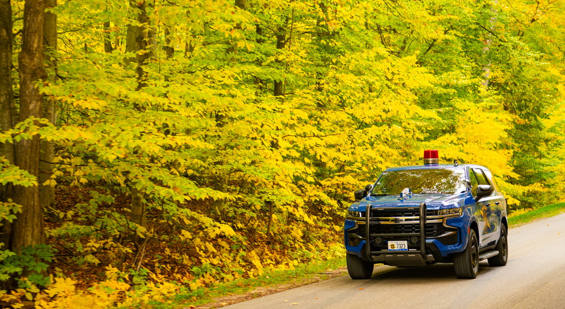 Blue Michigan State Police patrol car driving through forest with yellow leaves on trees. 