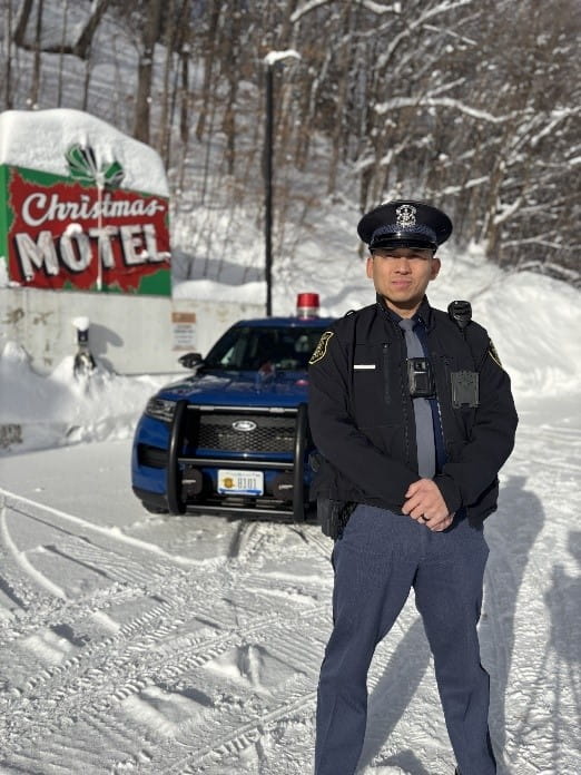 Male trooper standing outside of his patrol car with the Welcome to Christmas sign and snow in the background