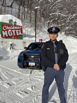 Male trooper standing outside of his patrol car with the Welcome to Christmas sign and snow in the background