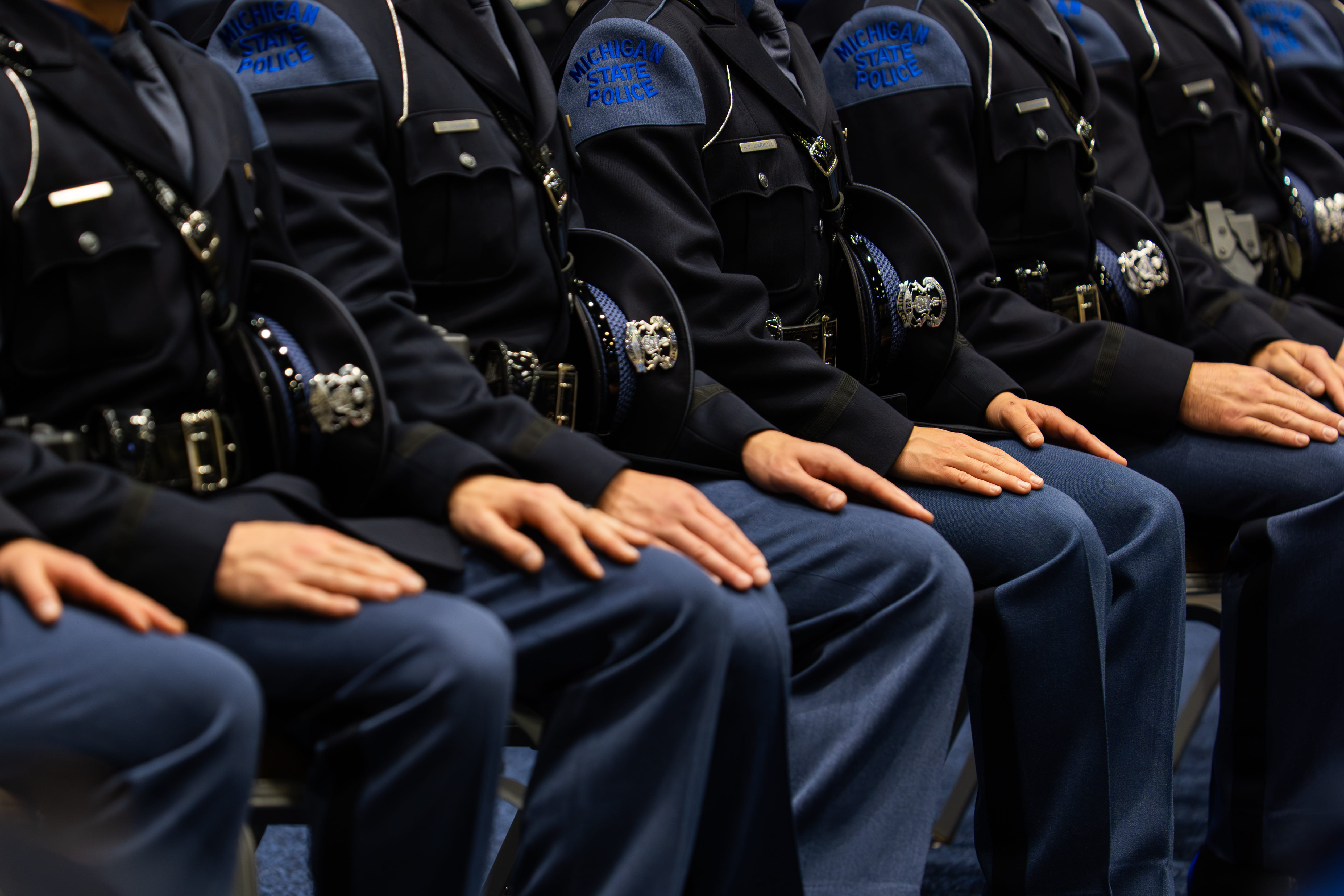 Troopers sitting in chairs during their graduation ceremony with hands and hats on their laps