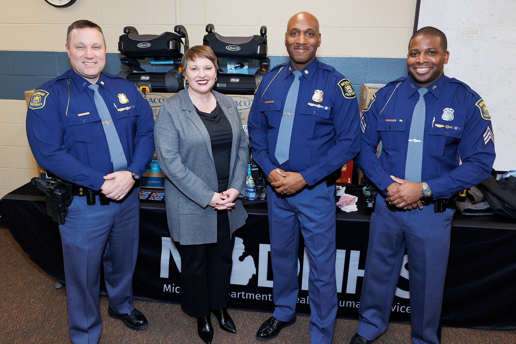 MSP Insp. Barry Schrader, MDHHS Director Elizabeth Hertel, MSP Col. James F. Grady II and Sgt. Ronnie Evans standing in front of a table filled with diapers, warm clothes, formula, car seats
