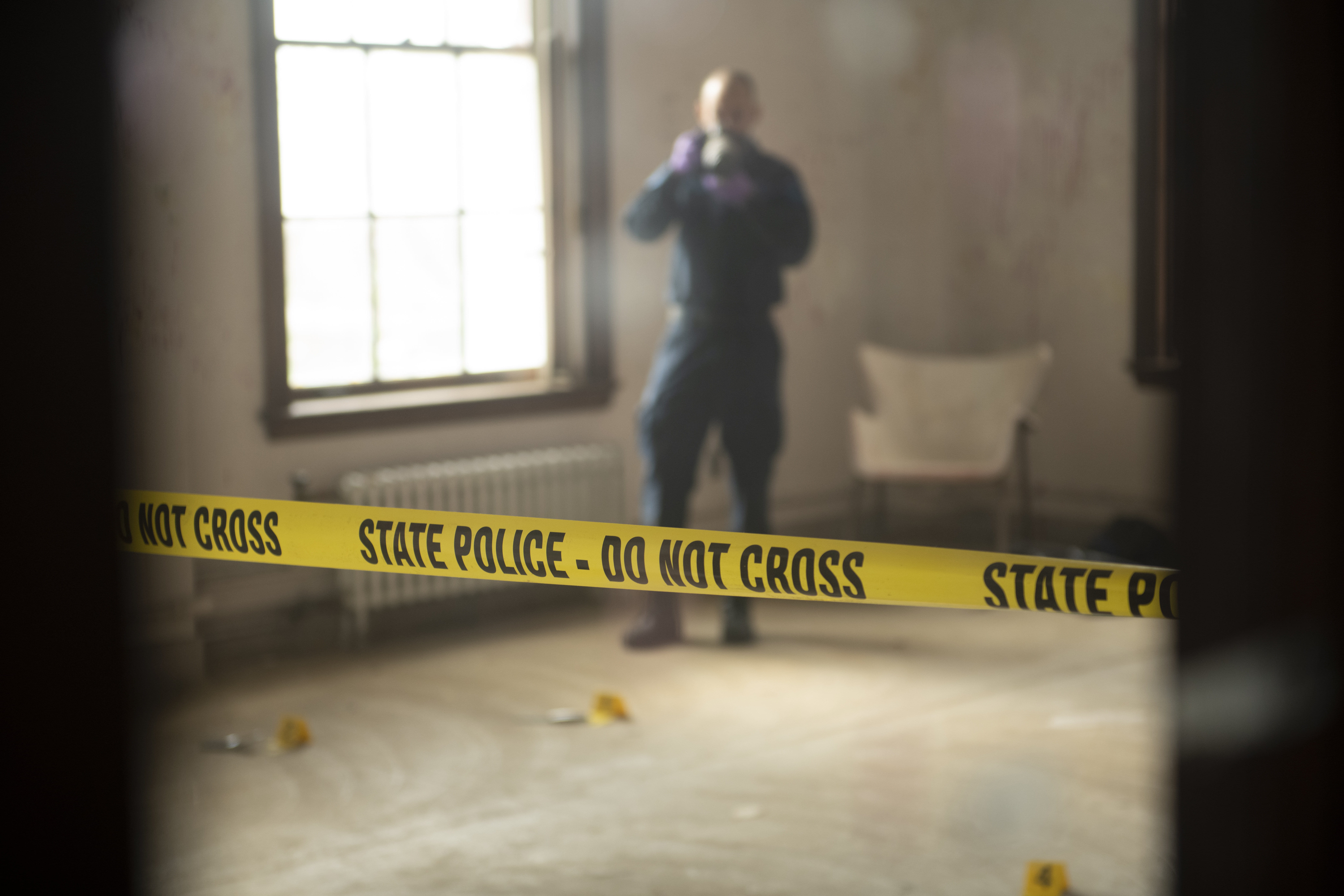 Crime scene tape in foreground with out-of-focus crime scene technician in background holding camera.