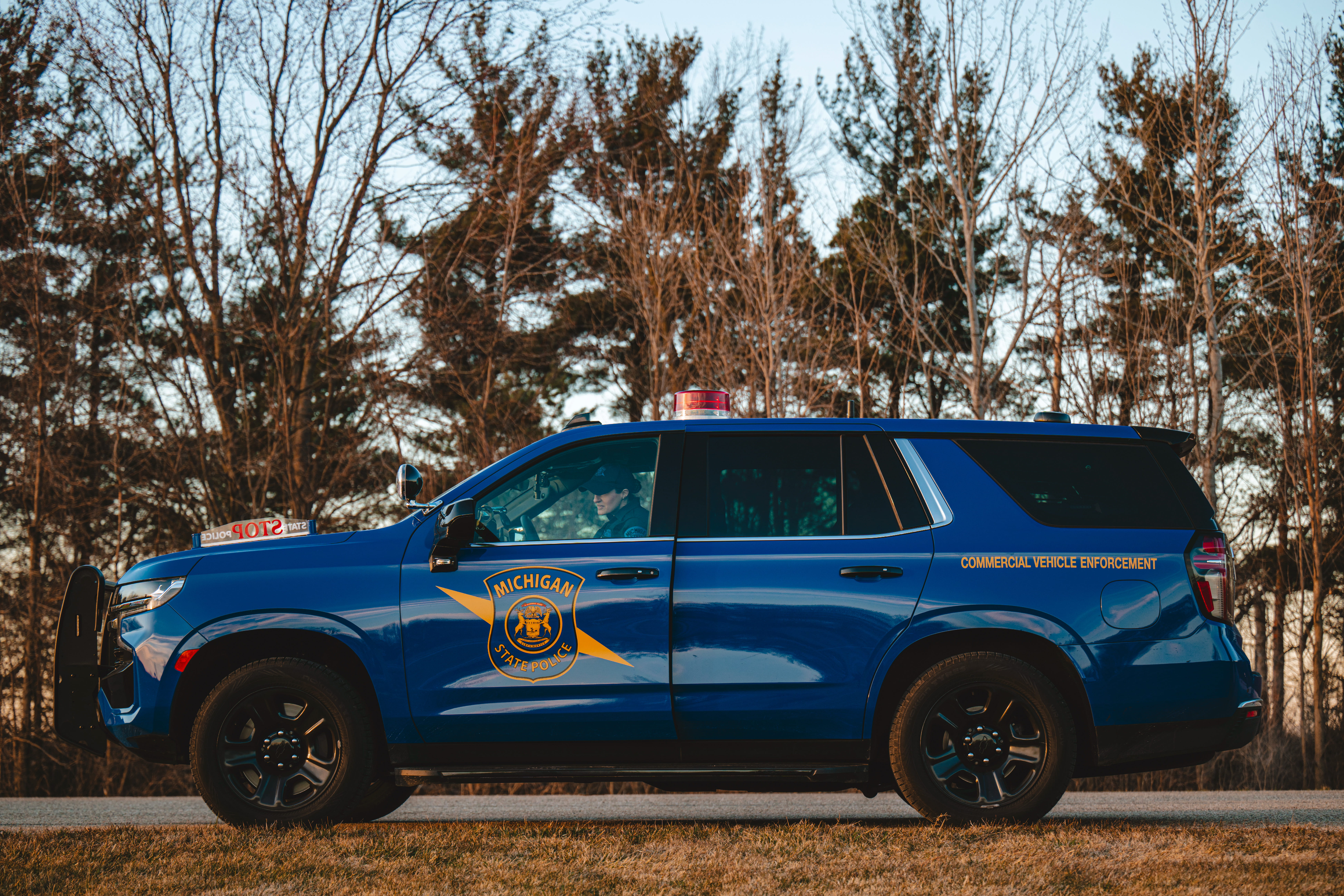 Michigan State Police Commercial Vehicle Enforcement patrol car in front of pine trees, with female officer at the wheel.