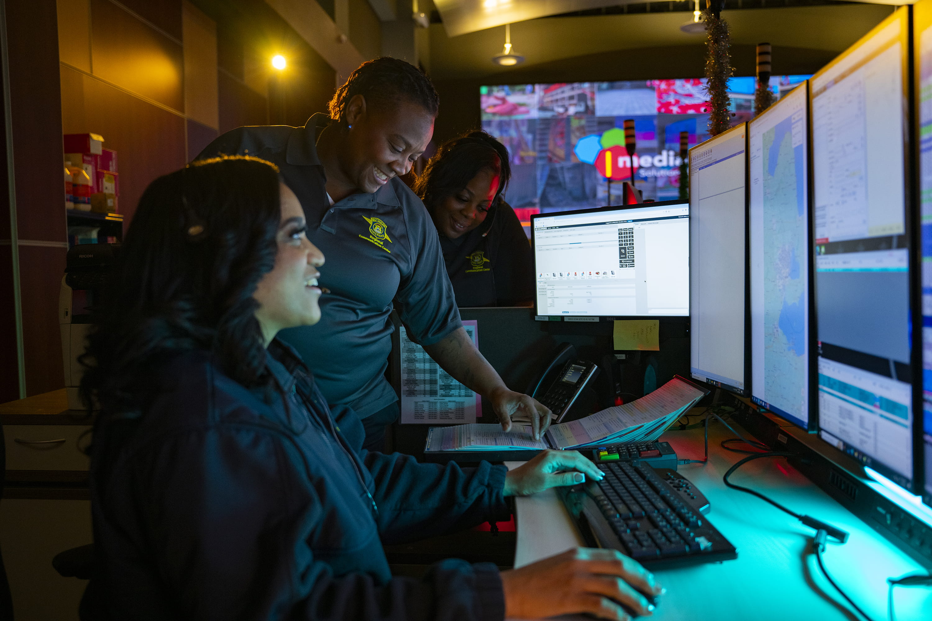 Emergency dispatcher wearing headset seated at a computer with two other dispatchers present in the background.