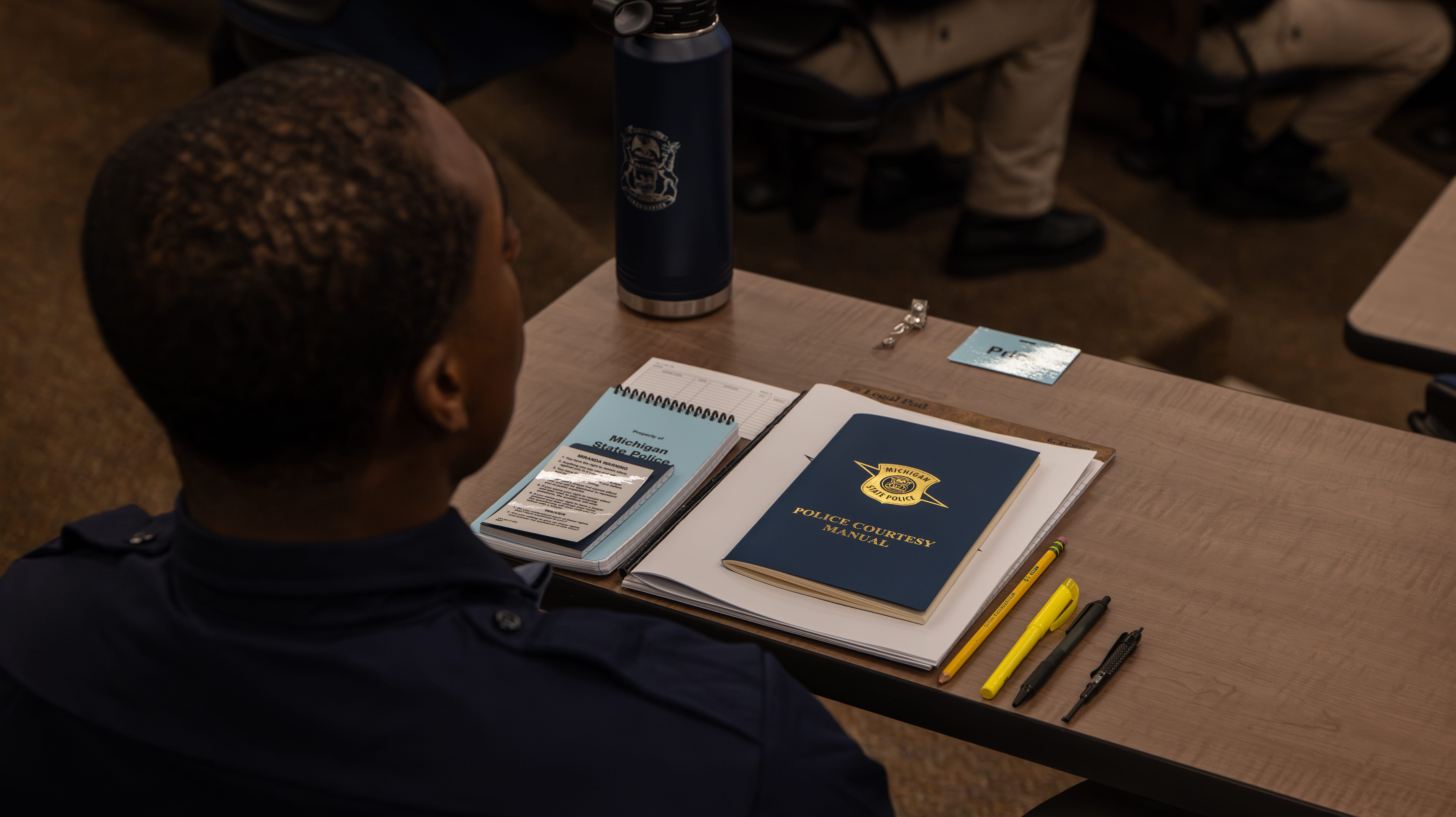 View over trooper recruit’s shoulder as he sits in auditorium with paper, police courtesy manual, and pens on desk in front of him.