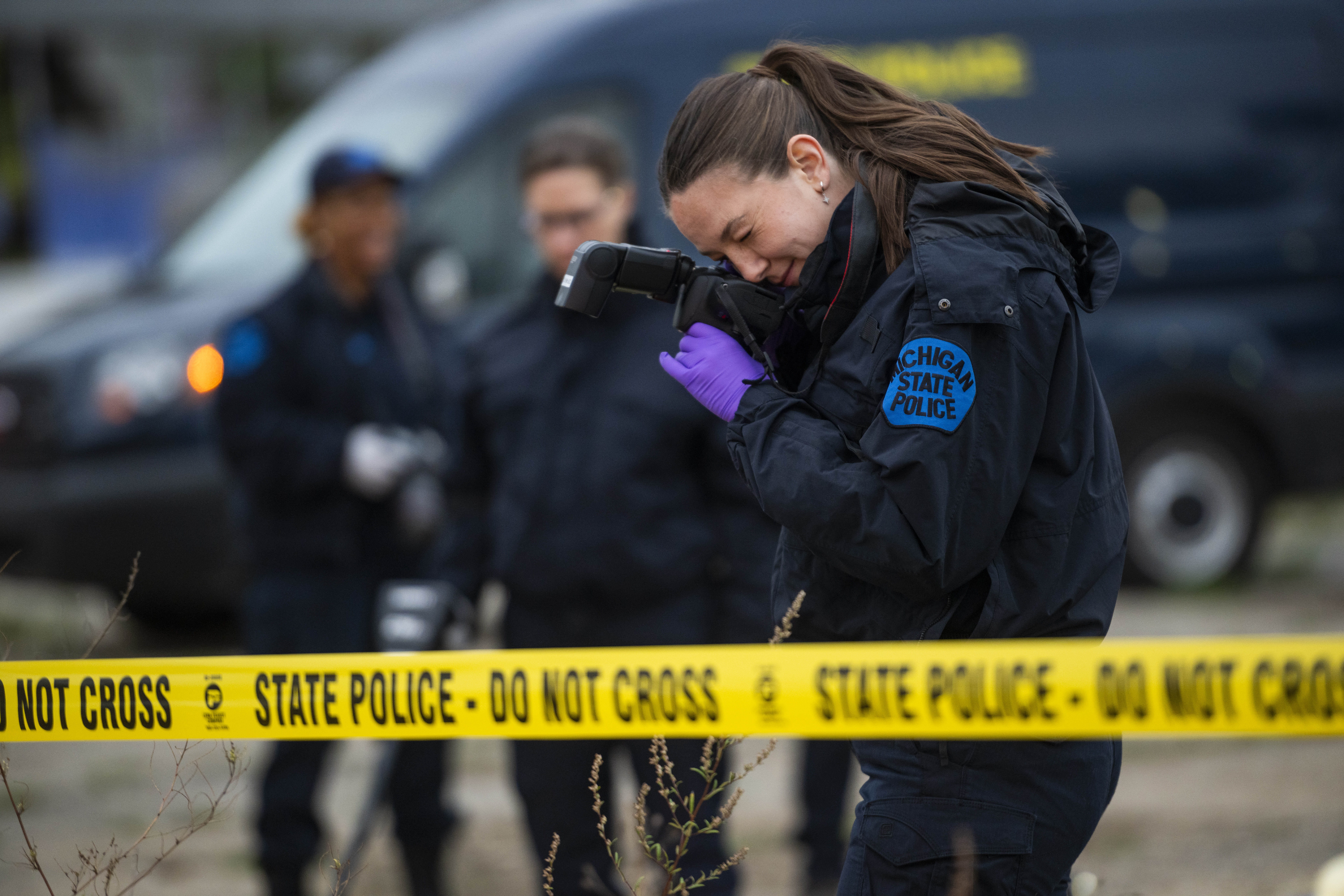 Crime scene tape and technician with camera in foreground, other technicians out-of-focus in background.