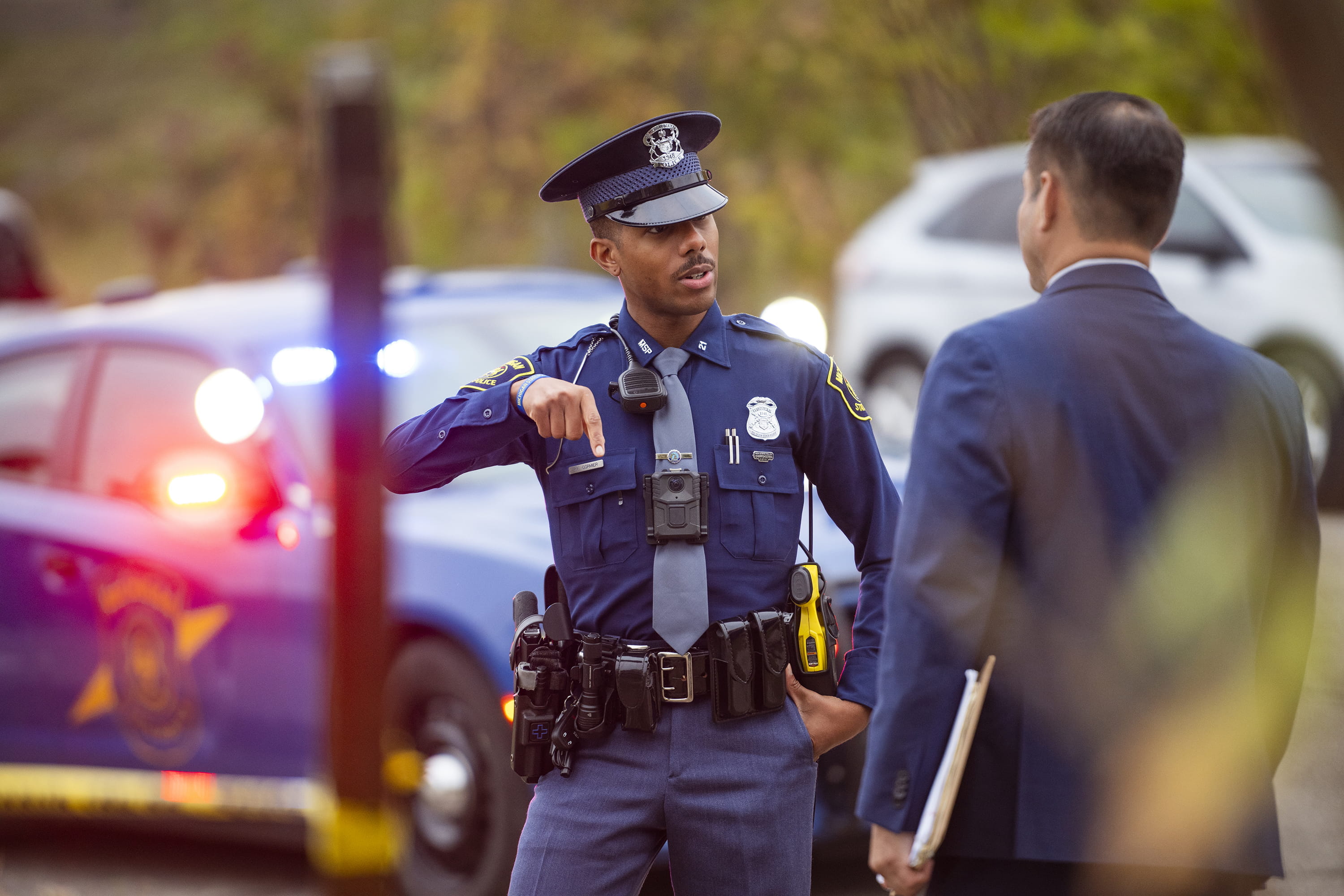 Trooper and detective in discussion, patrol car in background.