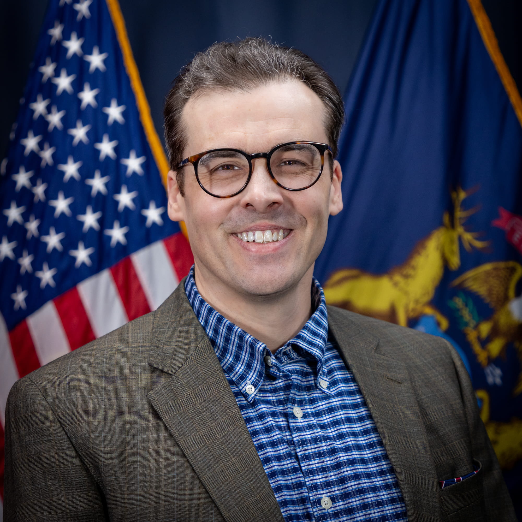 Zachary Dillinger wearing a blue shirt and tan jacket stands in front of the American and Michigan flags