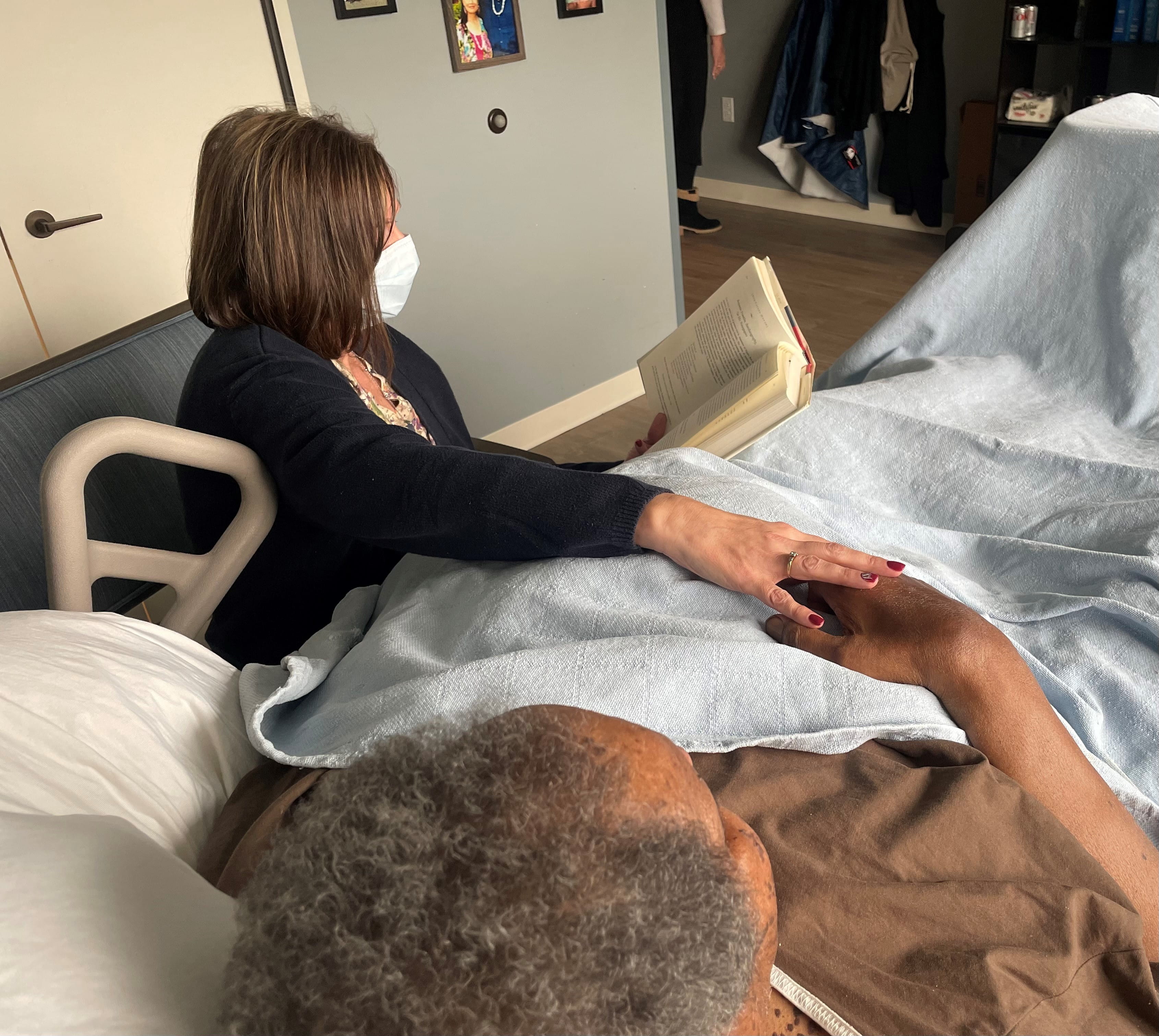 Woman sits bedside reading a book to a veteran