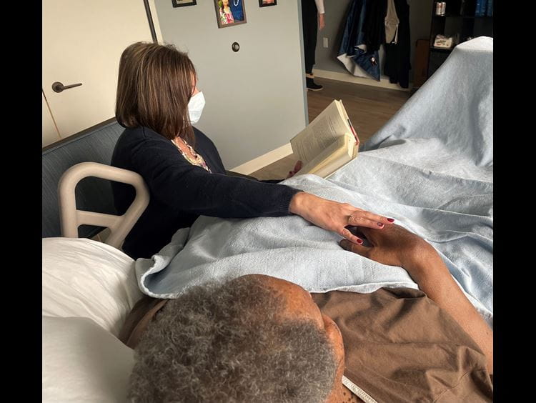 Woman sits bedside reading a book to a veteran
