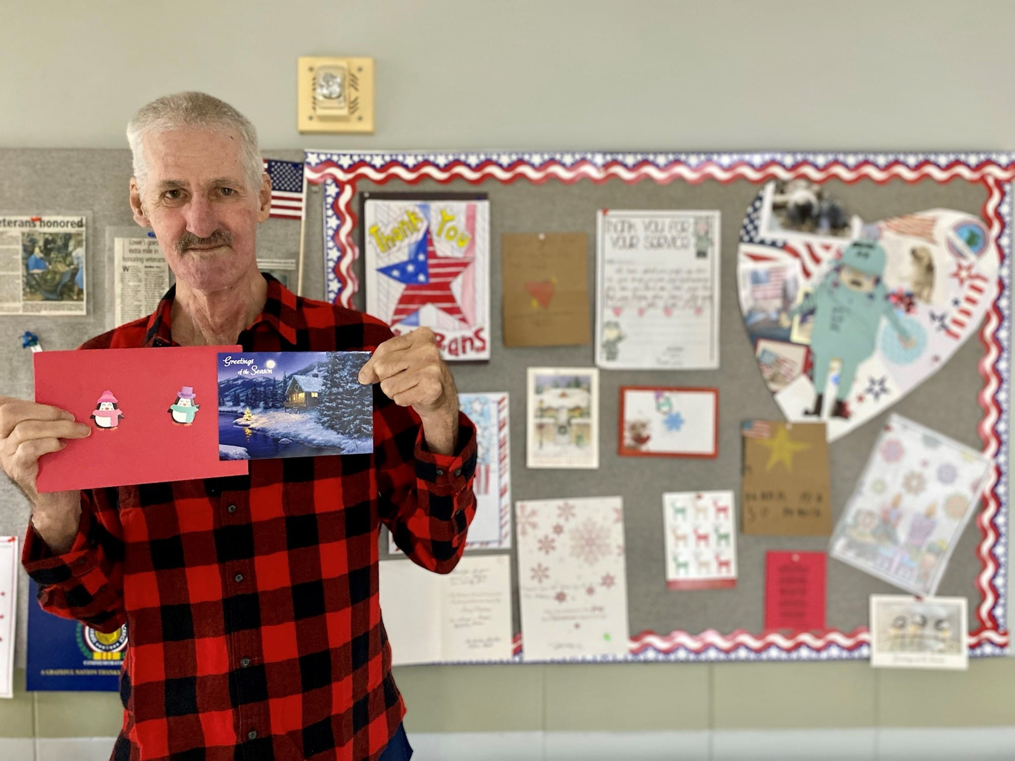 Veteran wearing a flannel shirt holds a holiday card in his hands with a bulletin board full of cards in the background