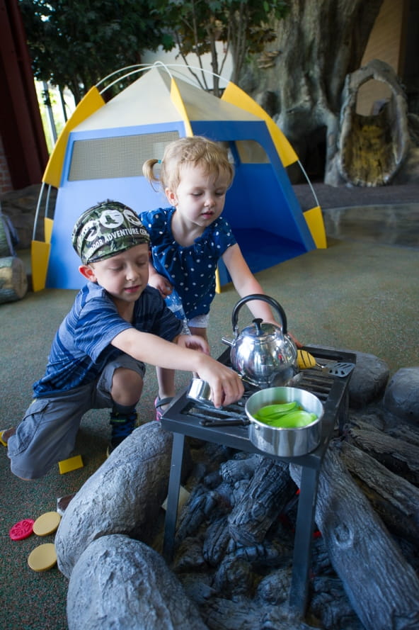 young boy and girl cooking pretend food over campfire in Outdoor Adventure Center campsite exhibit
