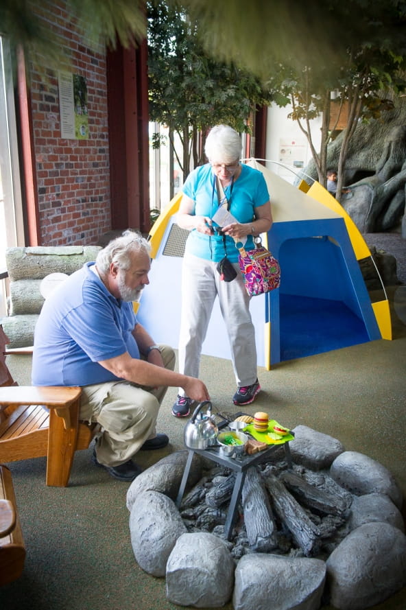 older couple in the Outdoor Adventure Center campsite, man sitting by campfire cooking pretend food