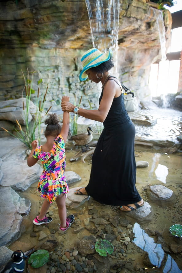 mom helps young girl cross rocks in pond at bottom of Outdoor Adventure Center waterfall