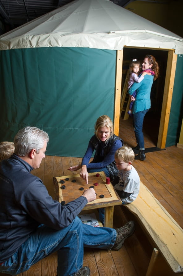 family plays checkers at a wooden table outside yurt replica at the Outdoor Adventure Center