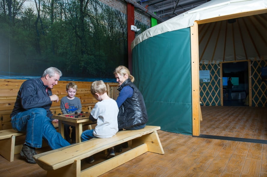 family plays checkers at a wooden table outside yurt replica at the Outdoor Adventure Center