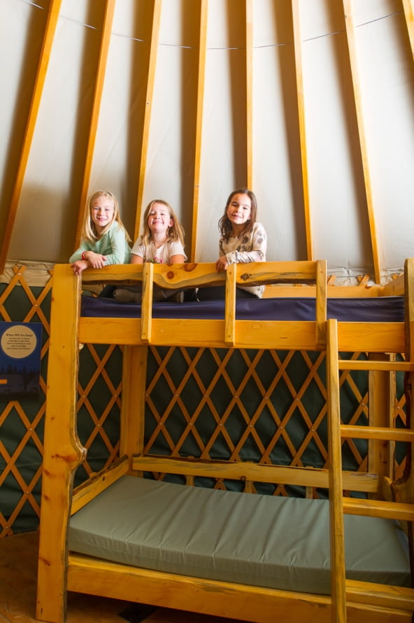 three young girls sit on top bunk of wooden bunkbed in yurt replica at the Outdoor Adventure Center