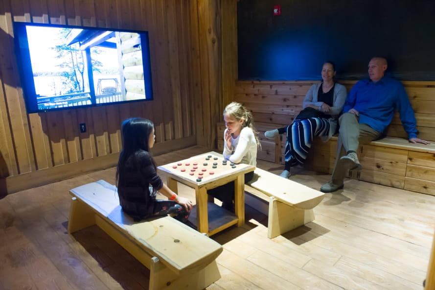 girls play checkers at a wooden table, with lodging scenes on a monitor, at Outdoor Adventure Center