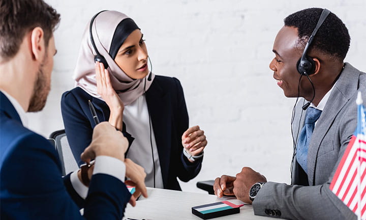 a woman in a hijab and a black man sitting at a table facing each other wearing headsets and practicing languages while man across table times them