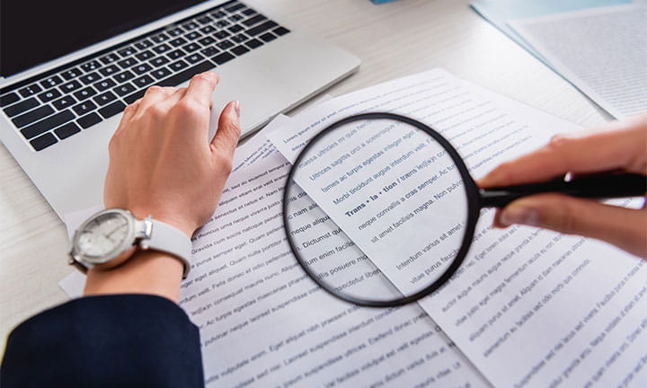 arms and hands of a person at a computer with translated papers in front of them holding a magnifying glass to the paper