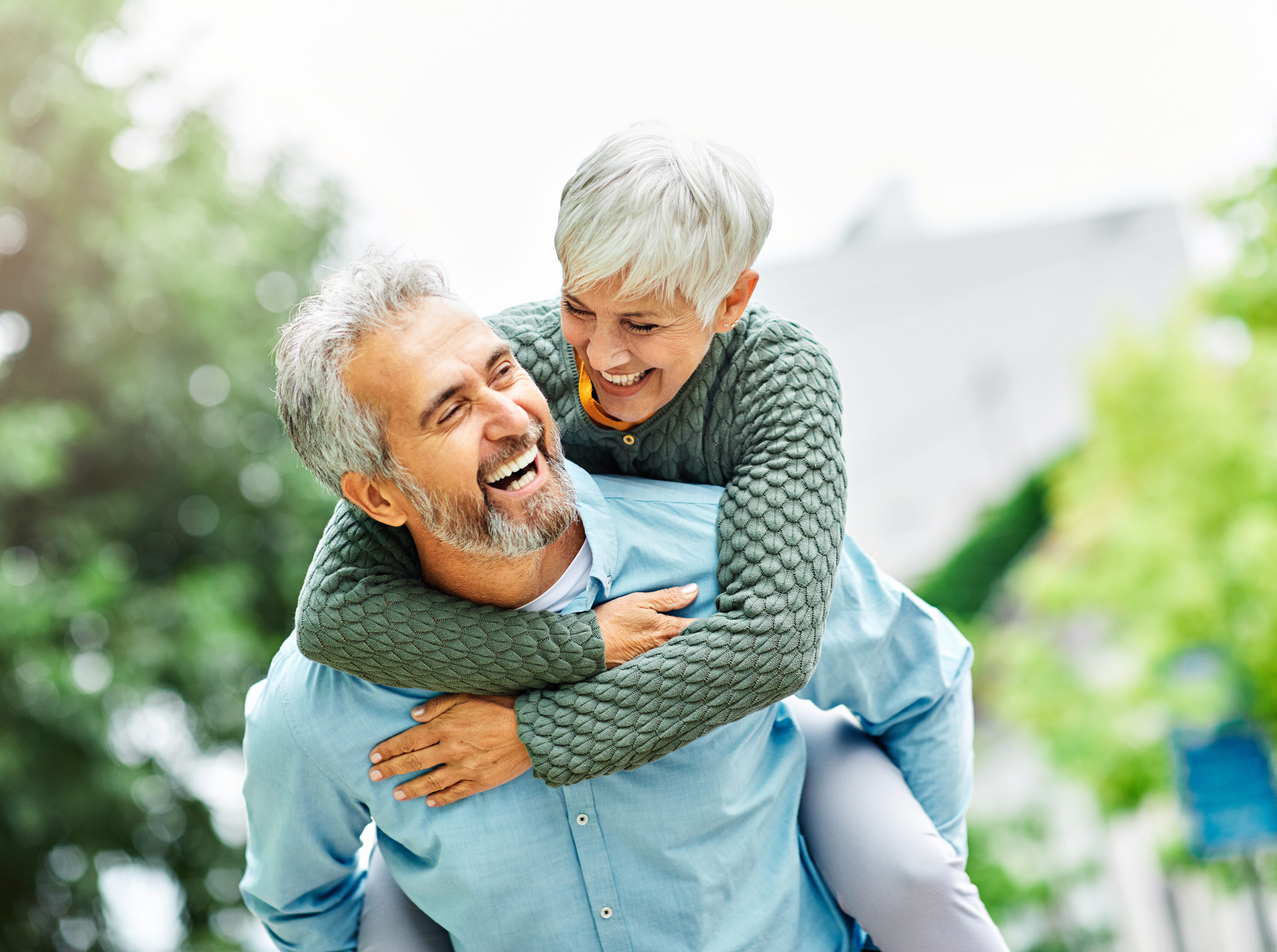 A laughing older White man gives a playful piggyback ride to a smiling older White woman with short silver hair outdoors. They both look joyful against a soft green background.