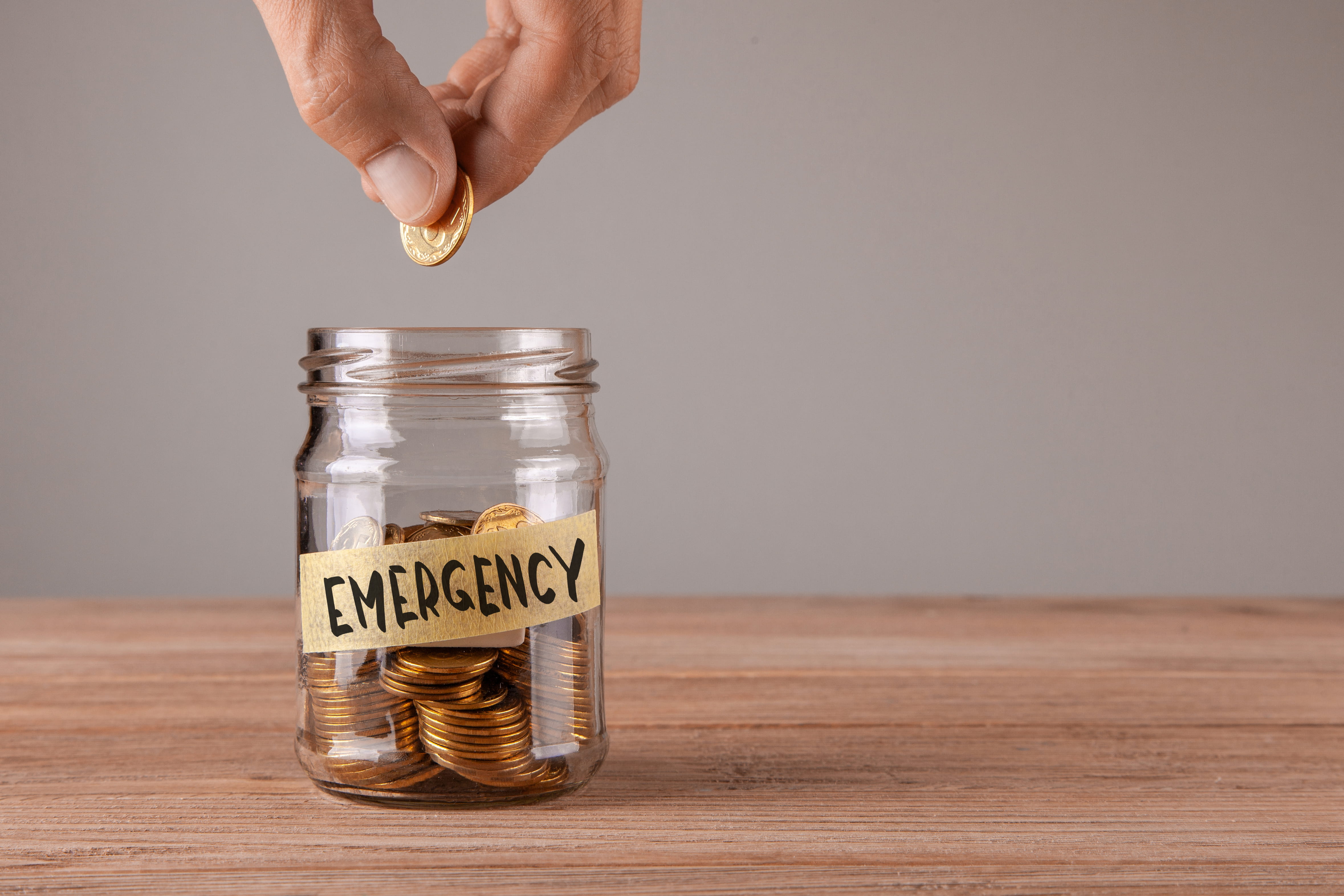 A hand drops a gold coin into a glass jar filled with coins. The jar has a label that reads "EMERGENCY" in black ink, sitting on a wooden surface against a neutral background.