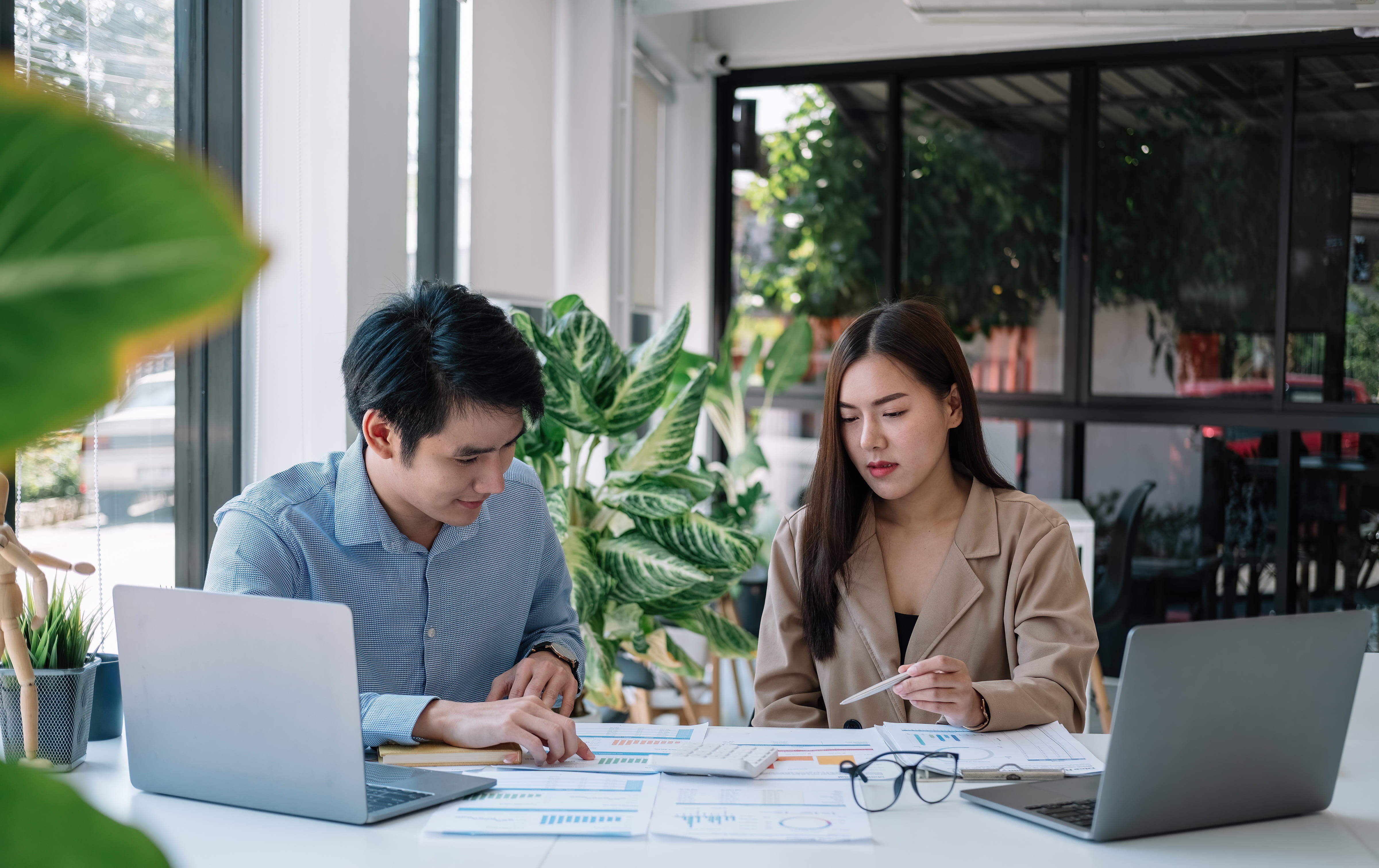 Two colleagues sit at a desk in a bright office, reviewing printed data charts and using laptops. A man points at a document while a woman holds a pen.