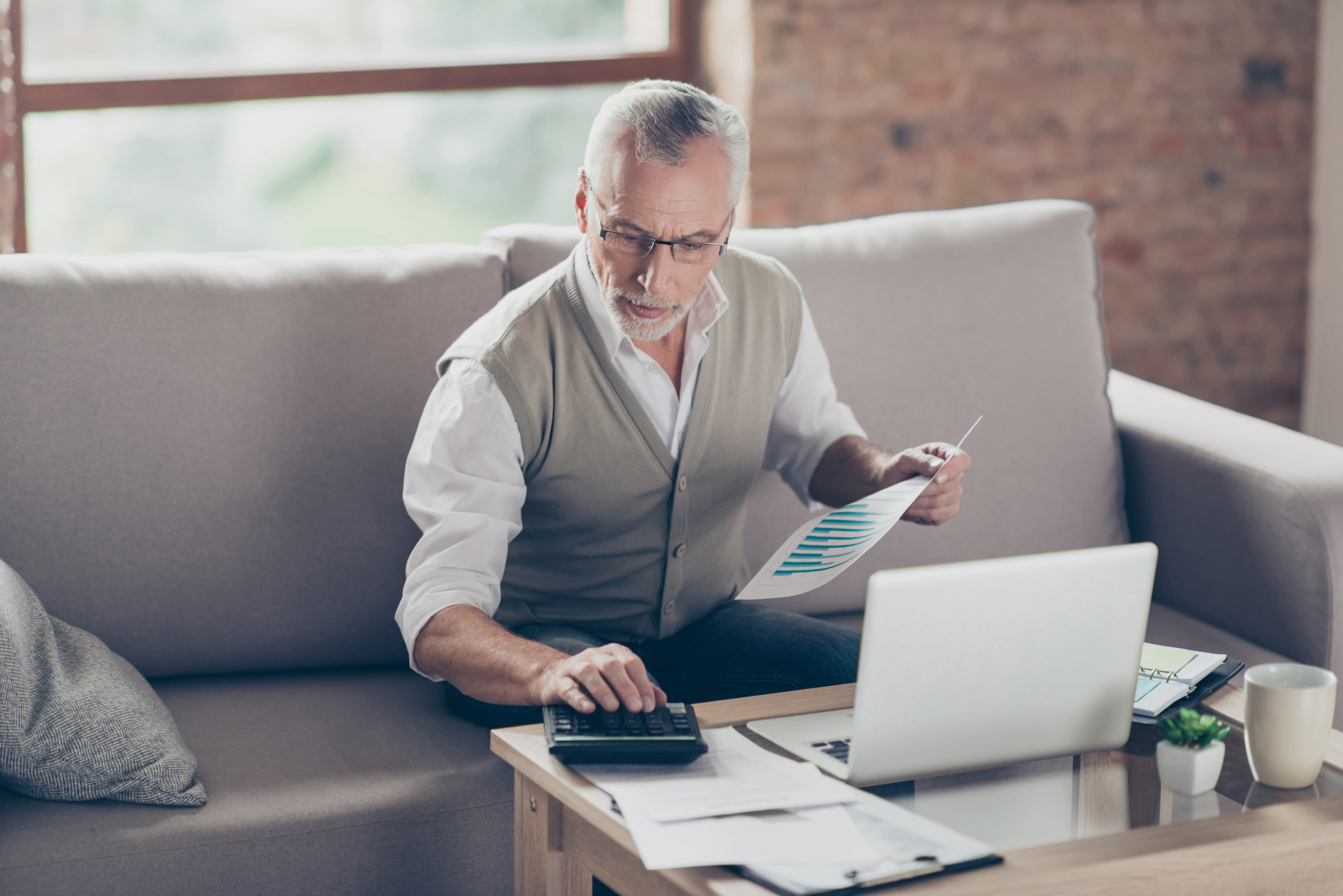 A white senior man in a sweater vest using a calculator and laptop to review a document with financial charts while sitting on a couch.