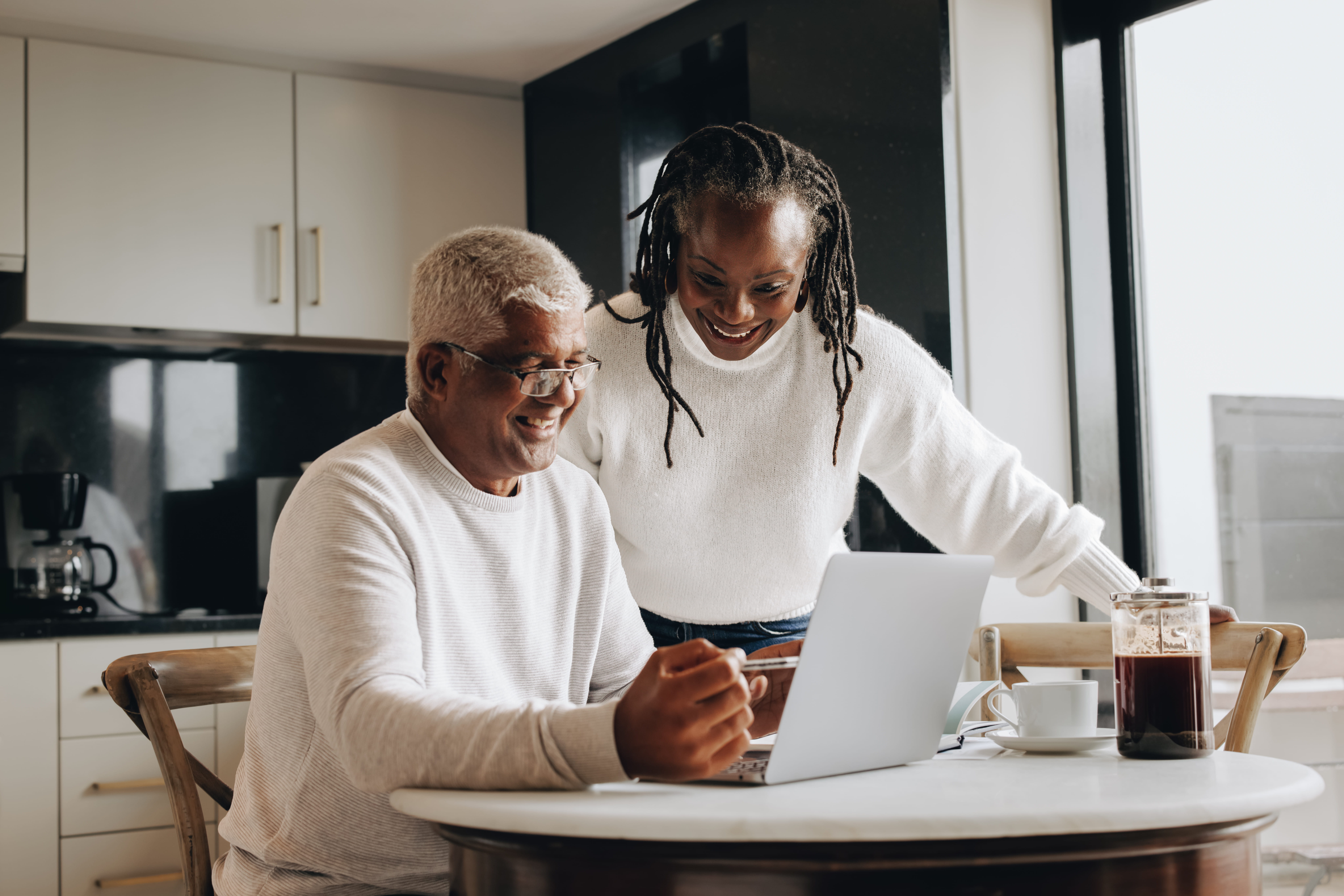 A smiling Black couple in a bright kitchen looking at a laptop together; the man is seated while the woman leans in from behind.
