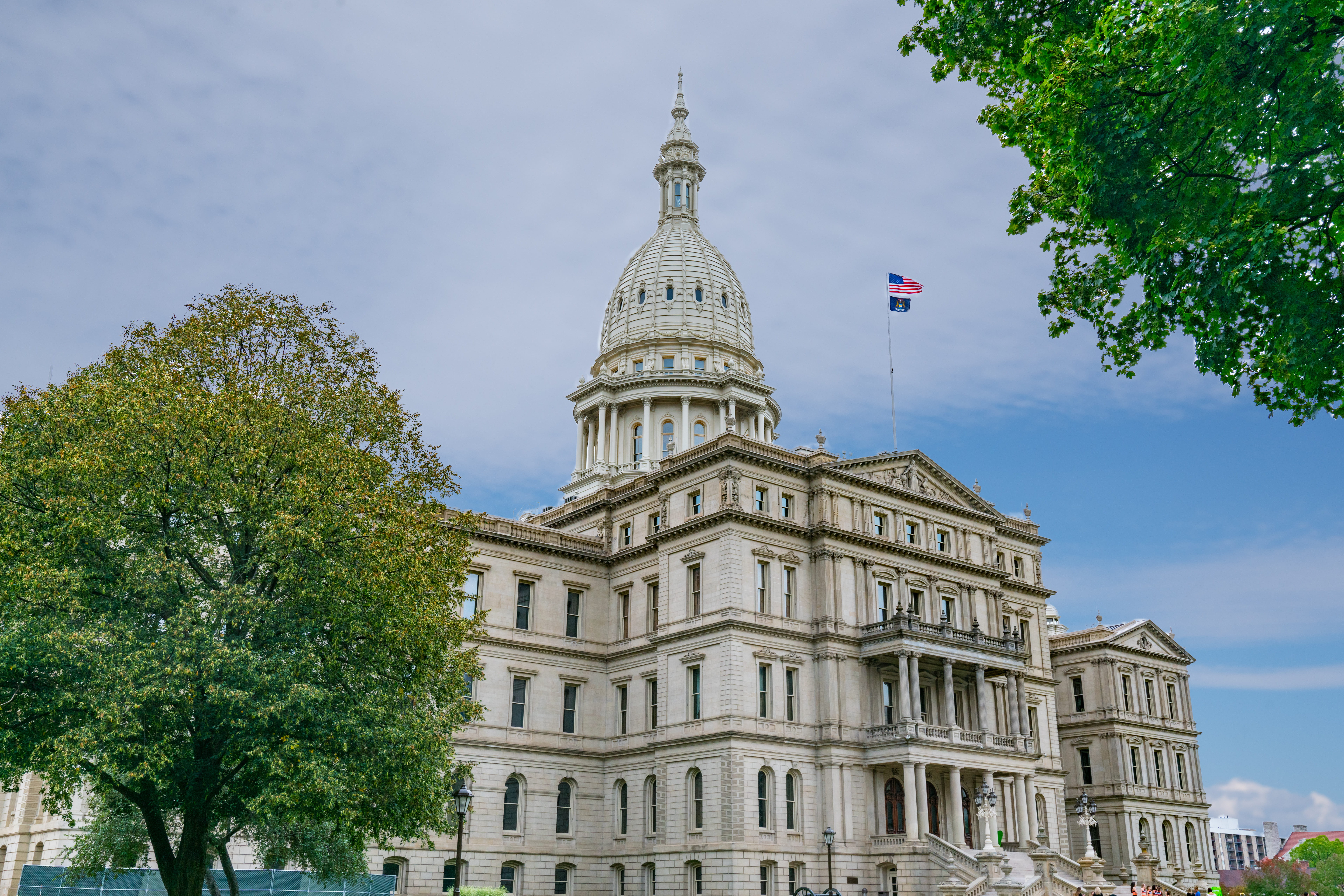 The Michigan State Capitol building in Lansing, featuring its white neoclassical dome and limestone facade under a slightly cloudy blue sky, with the American and Michigan state flags flying.