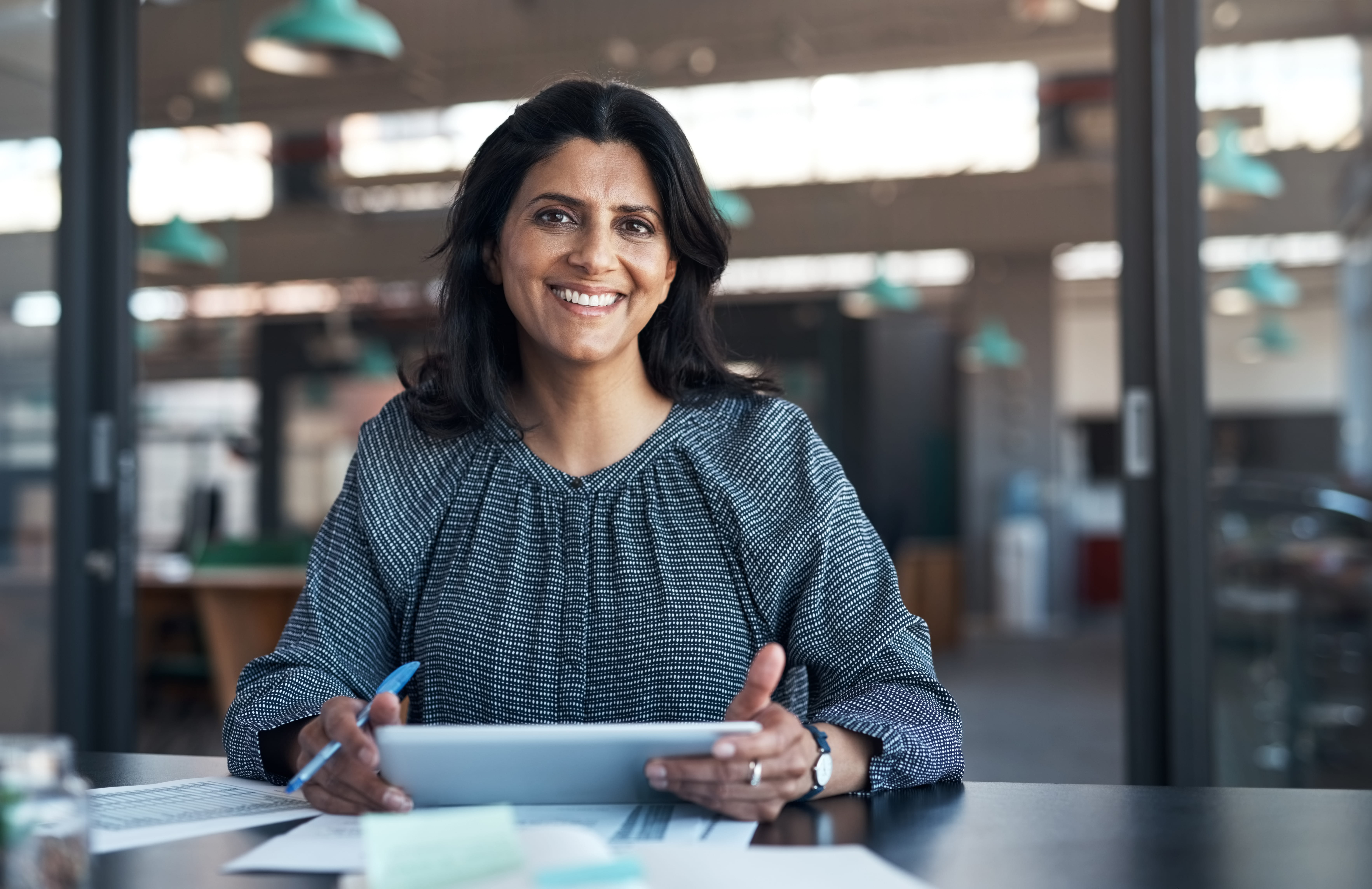 A smiling woman with medium-toned skin and dark hair sits at a desk in a modern office, holding a digital tablet with documents spread out.