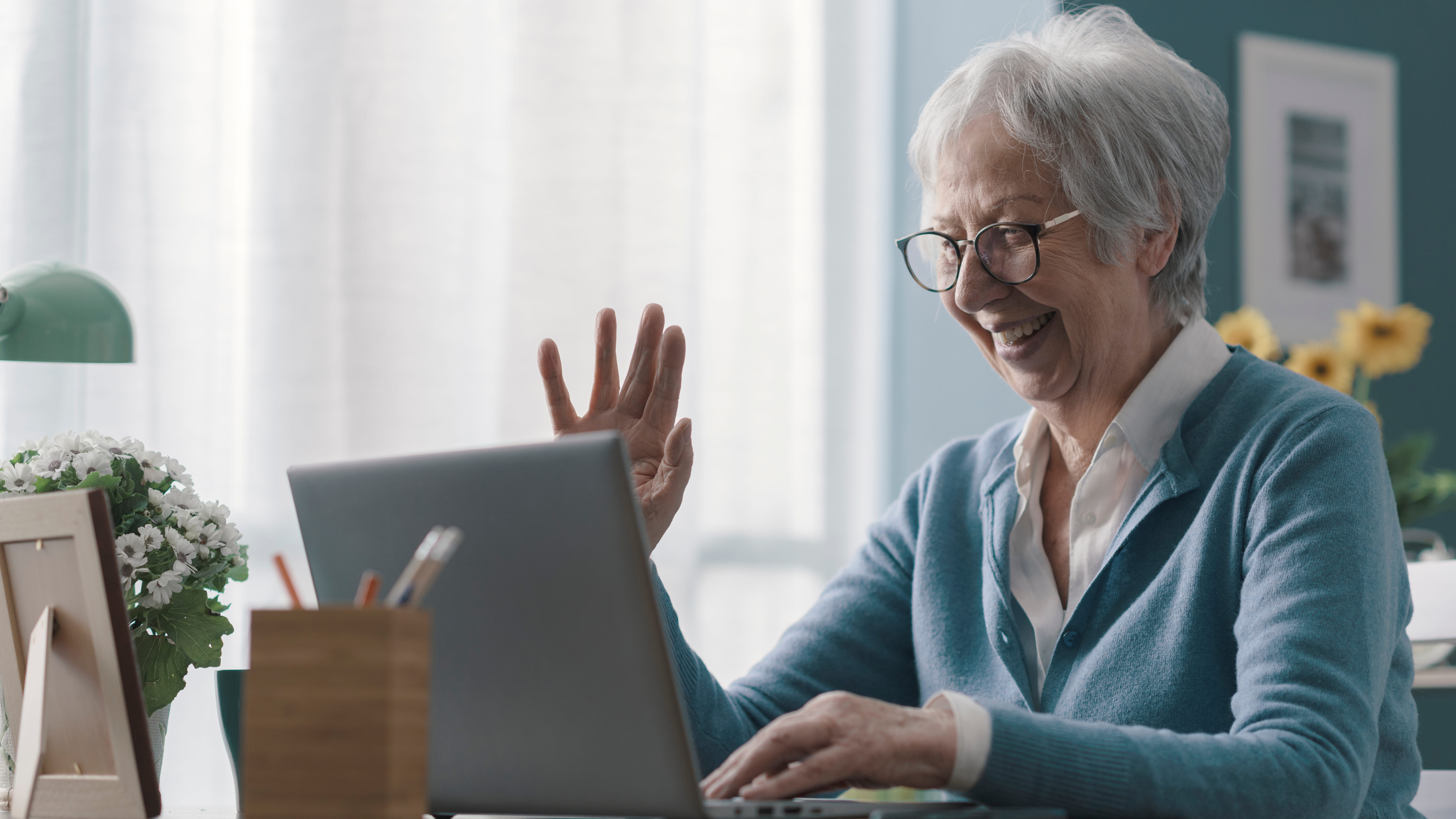 A senior white woman with short gray hair and glasses smiles and waves at her laptop screen while sitting at a desk with flowers.