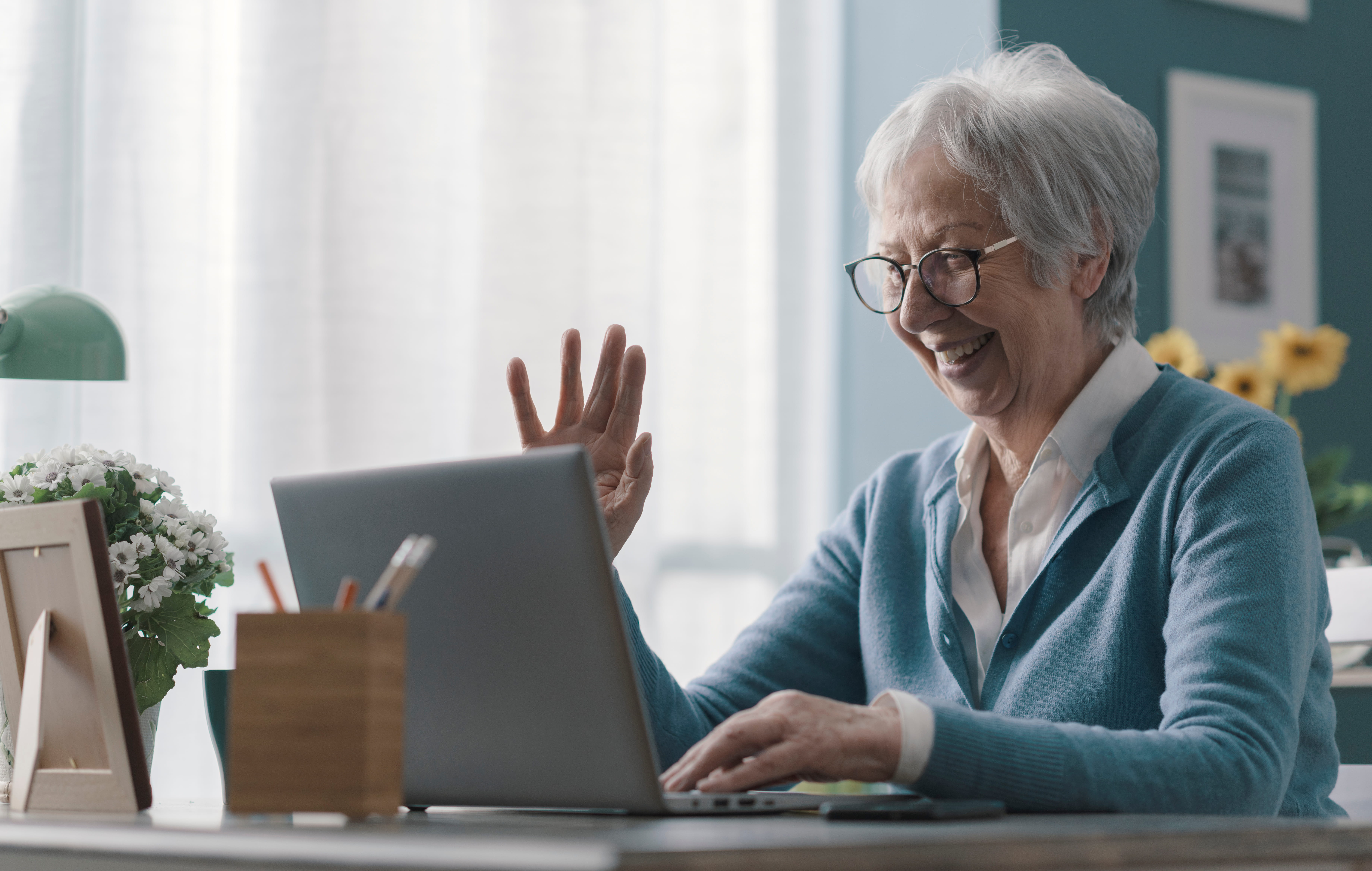 A senior white woman with short gray hair and glasses smiles and waves at her laptop screen while sitting at a desk with flowers.