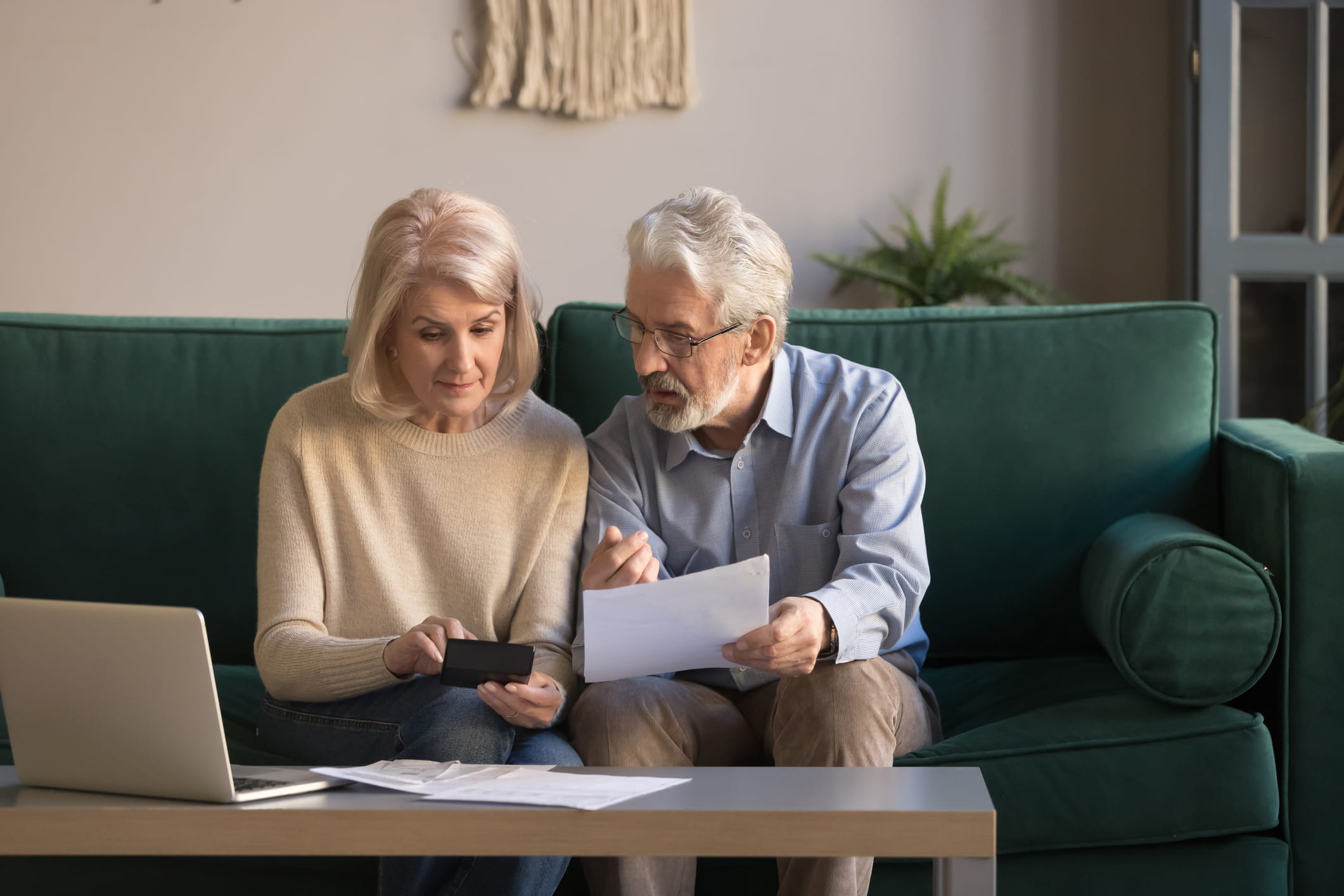 An older couple sits on a green sofa, focused on a document and a handheld calculator. A laptop and more papers are on the coffee table.
