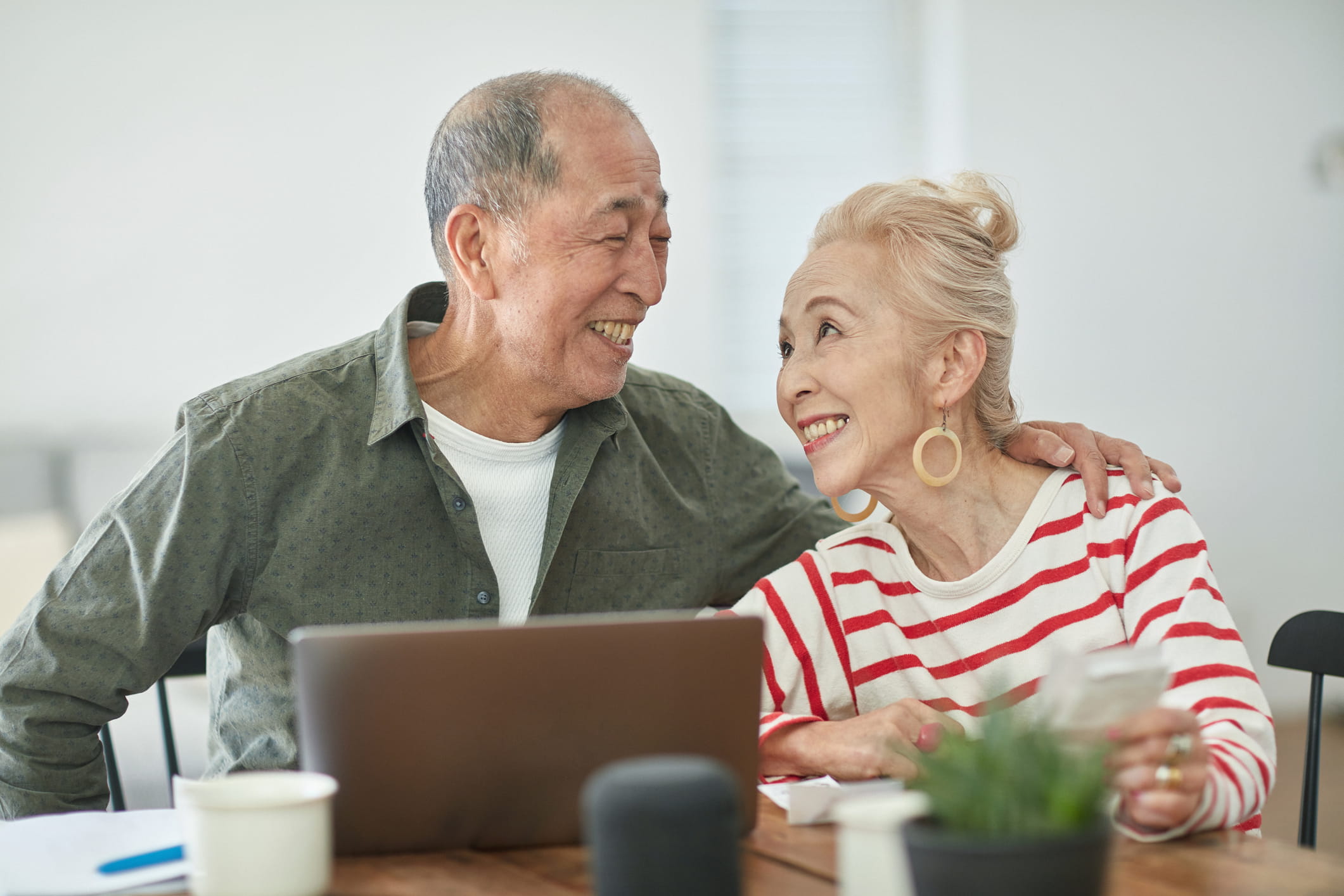 An older Asian couple smiles at each other while sitting at a table with a laptop and papers. They appear happy and relaxed.